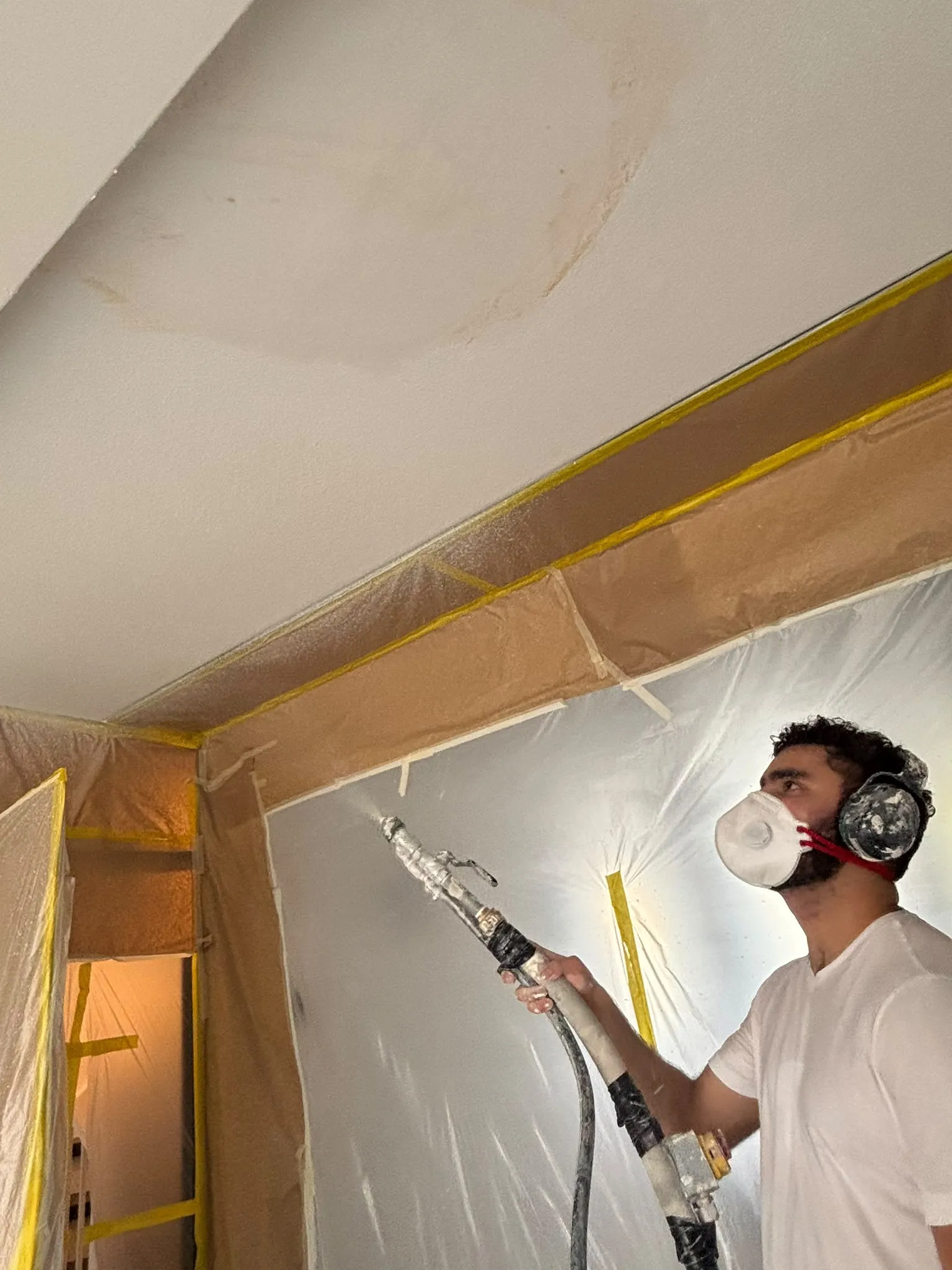 Stukadoor met mondkapje en gehoorbescherming spuit een plafond, wanden afgeschermd met folie en tape